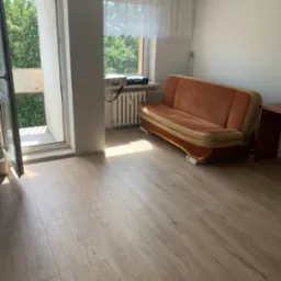 Pokój z panelami podłogowymi w odcieniach brązu, częściowo umeblowany: kanapa na kartonach i biurko, widok na balkon i zieleń za oknem.