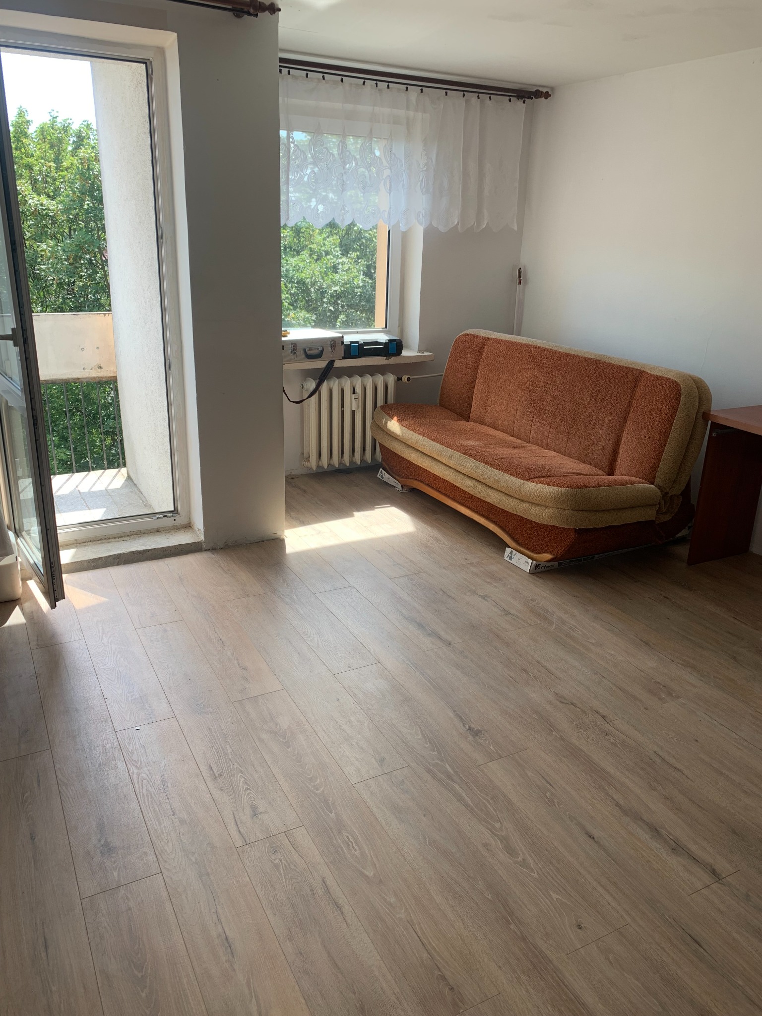 Pokój z panelami podłogowymi w odcieniach brązu, częściowo umeblowany: kanapa na kartonach i biurko, widok na balkon i zieleń za oknem.