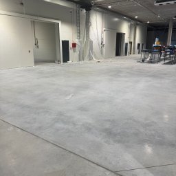 GLOBAL.ELEKTRO Patryk Piotrowski - Wnętrze hali produkcyjnej z widocznymi przewodami elektrycznymi pod sufitem, instalacjami przy ścianach i oświetleniem. Surowy beton na podłodze.