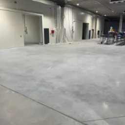 Wnętrze hali produkcyjnej z widocznymi przewodami elektrycznymi pod sufitem, instalacjami przy ścianach i oświetleniem. Surowy beton na podłodze.