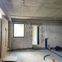 GLOBAL.ELEKTRO Patryk Piotrowski - Wnętrze w stanie surowym z widoczną instalacją elektryczną w peszlach, szarą skrzynką rozdzielczą na ścianie z bloczków i fragmentem okna z czarną ramą.