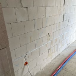 GLOBAL.ELEKTRO Patryk Piotrowski - Ściana z betonu komórkowego z zamontowanymi białymi listwami kablowymi i wystającymi przewodami. Widoczne rury w izolacji czerwonej i niebieskiej.