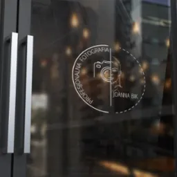 Grafika z minimalistycznym logo fotografa Joanny Bik, naklejona na szklane drzwi wejściowe do biura, widoczne srebrne uchwyty i odbicie miejskiego krajobrazu.