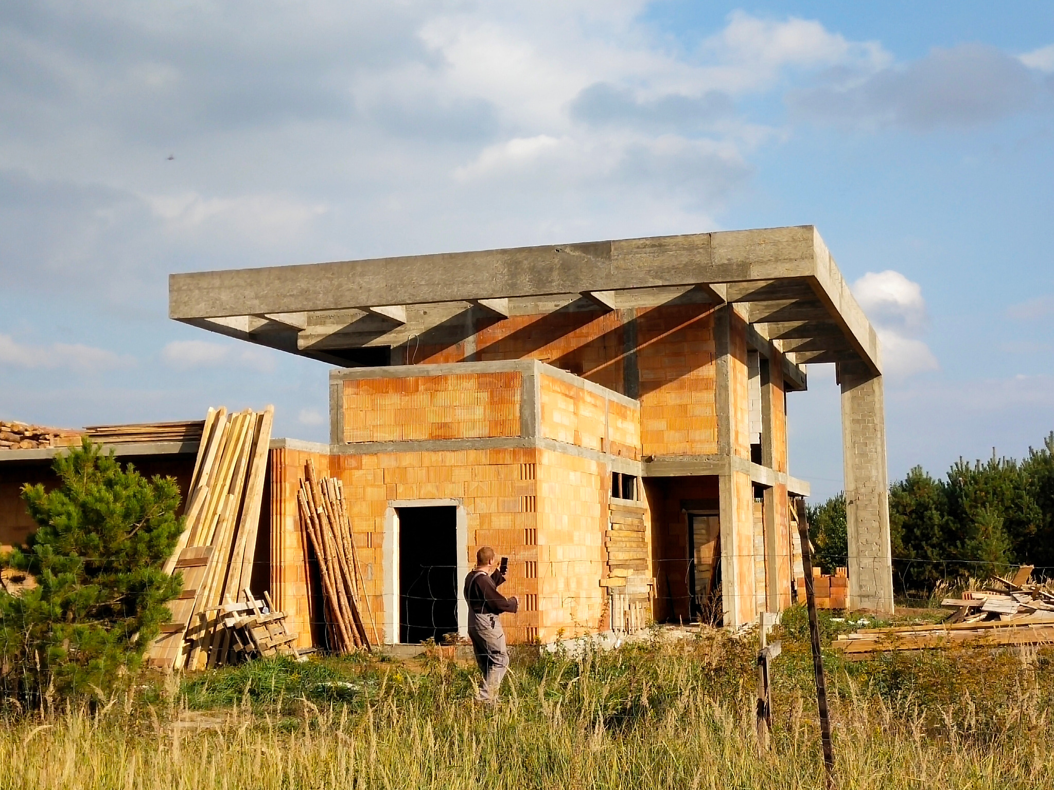 Niedokończony dwukondygnacyjny dom z pomarańczowej cegły z betonowym zadaszeniem, widoczny pracownik budowlany z telefonem, stos drewna i wysoka trawa.