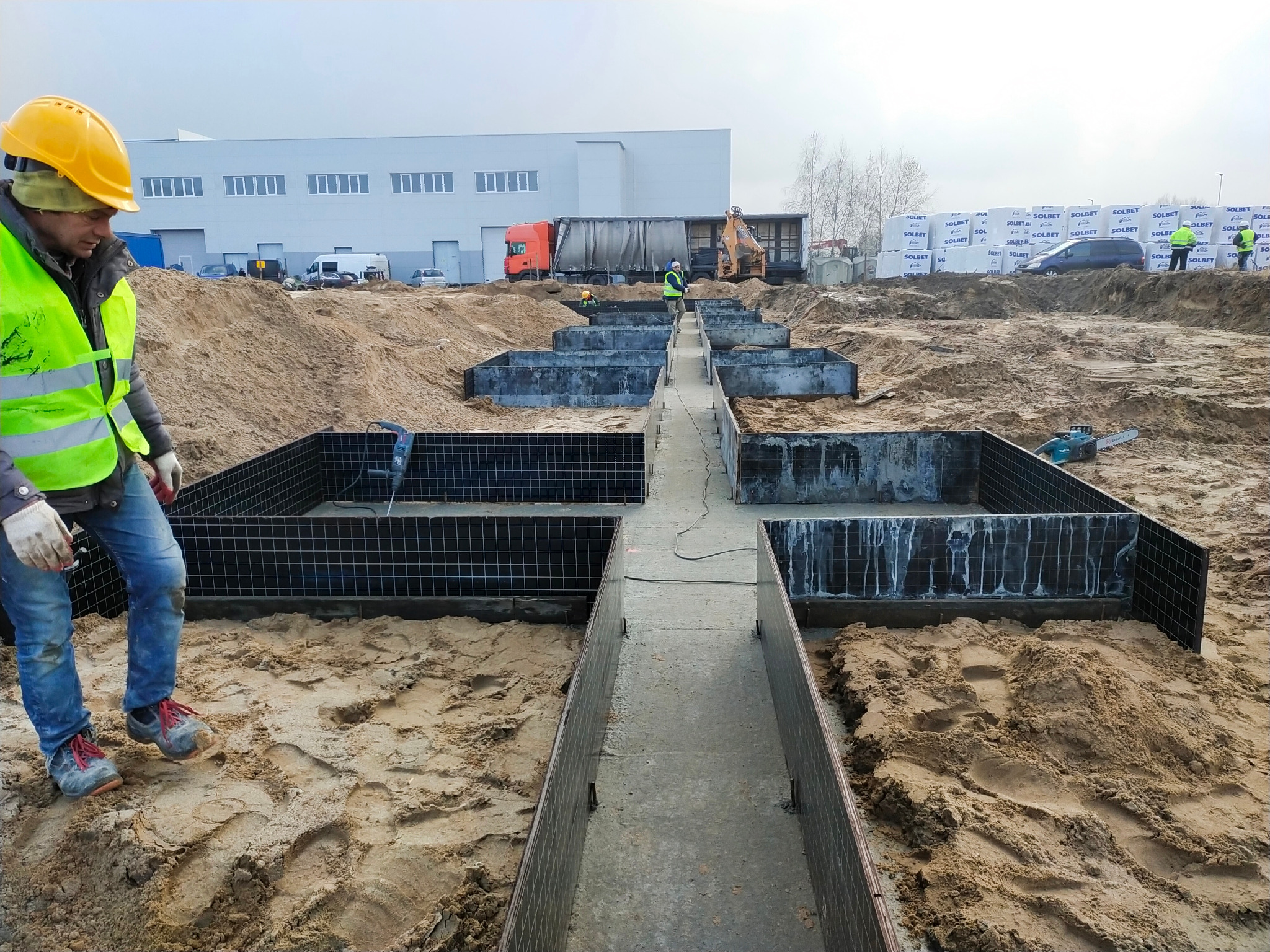 Przygotowanie fundamentów pod budynek: szalunki zbrojeniowe w wykopie, pracownik w żółtym kasku i odblaskowej kamizelce, w tle budynek przemysłowy i palety bloczków Solbet.