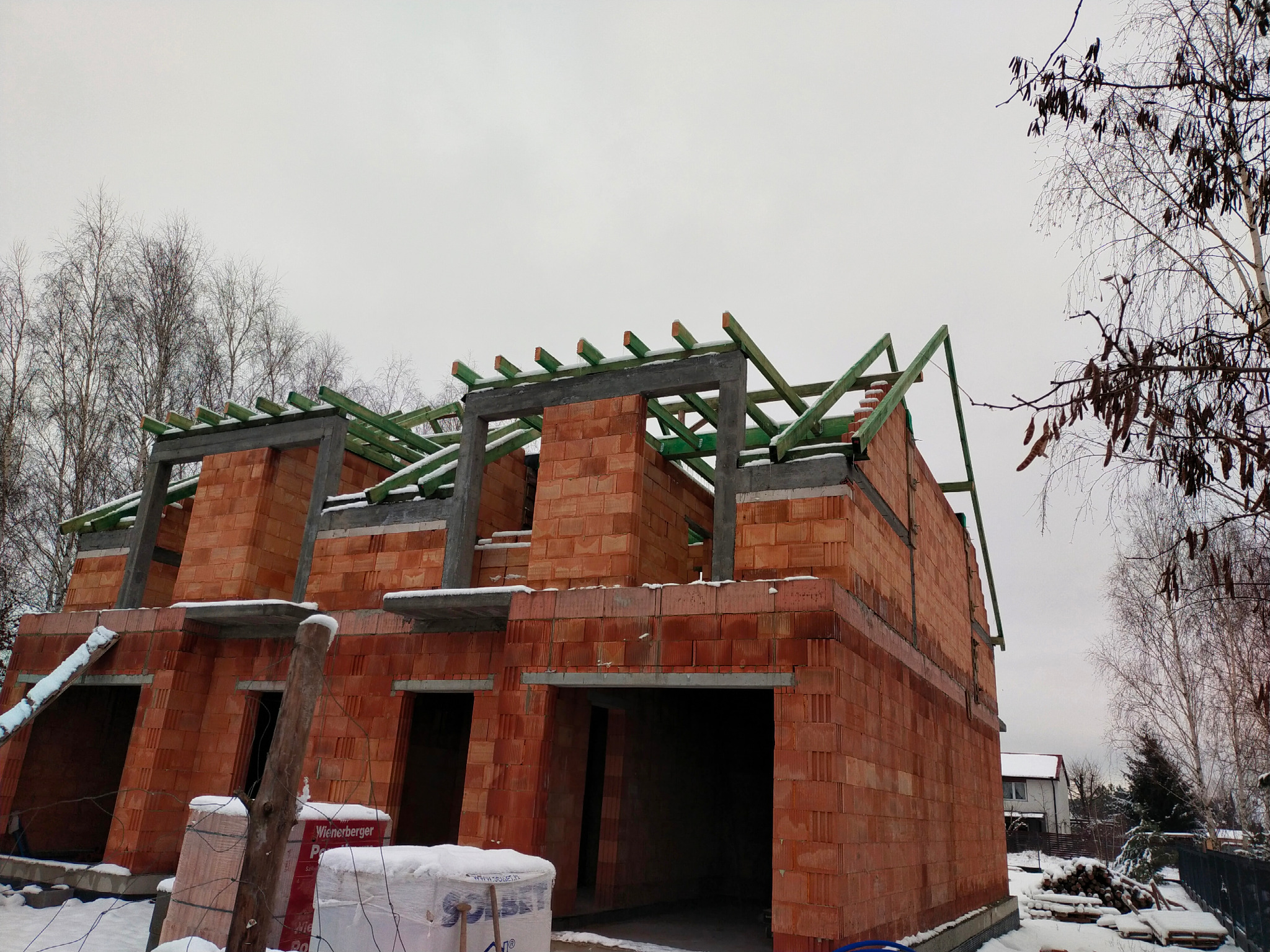 Wznoszenie ścian domu z czerwonej cegły, widoczne otwory okienne i drzwiowe, częściowo zadaszone zielonymi belkami, zimowy krajobraz z drzewami w tle.