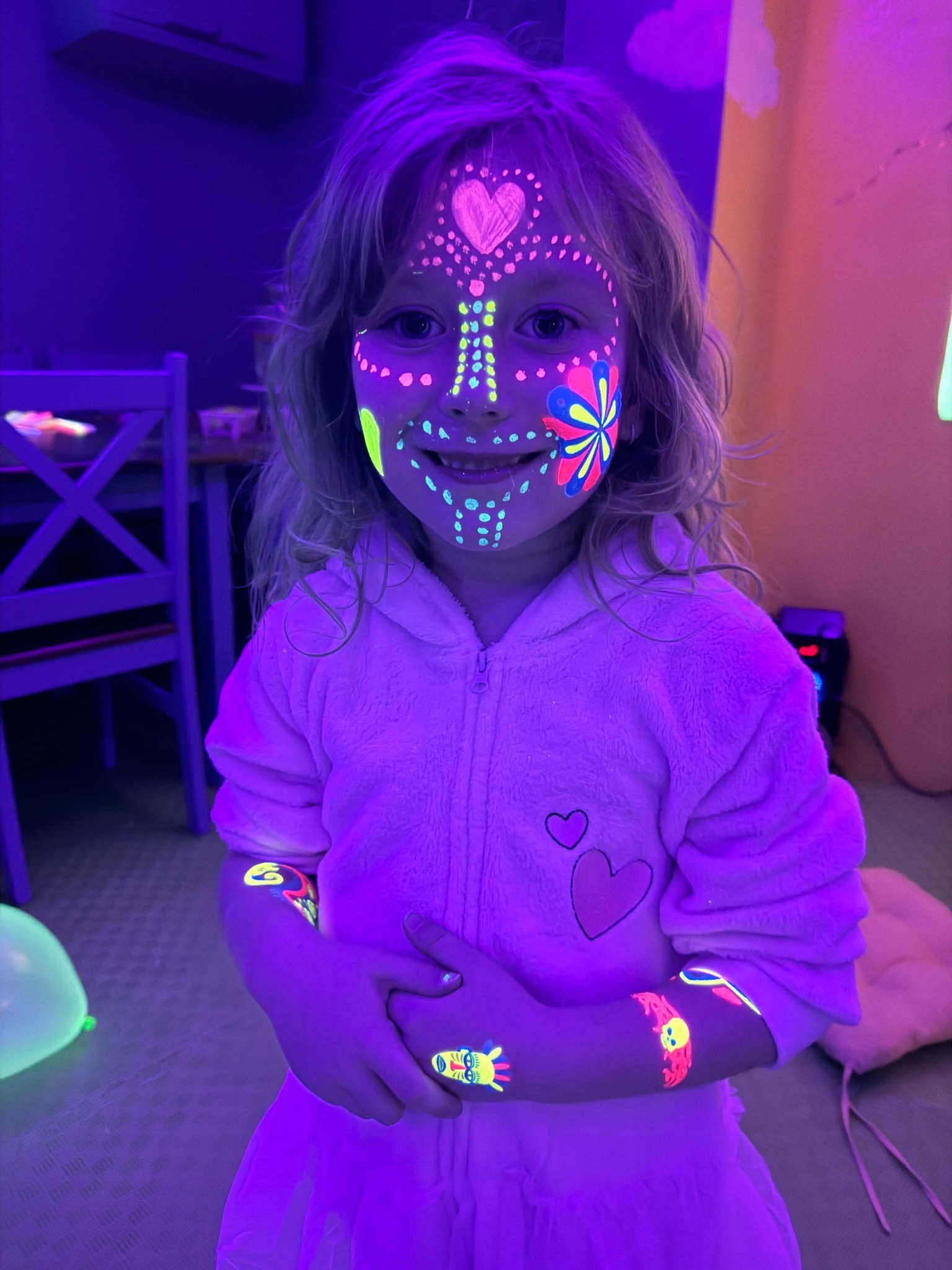 Uśmiechnięta dziewczynka z pomalowaną twarzą i ramionami farbami UV w ciemnym pomieszczeniu, widoczne elementy dekoracji imprezowej.