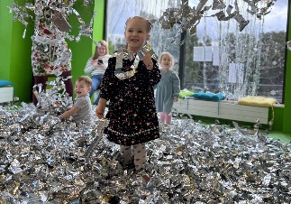 Dziewczynka w kwiecistej sukience uśmiecha się, stojąc po kolana w srebrnych, pogniecionych foliach podczas zabawy z innymi dziećmi w pomieszczeniu z zielonymi ścianami i dużym oknem.