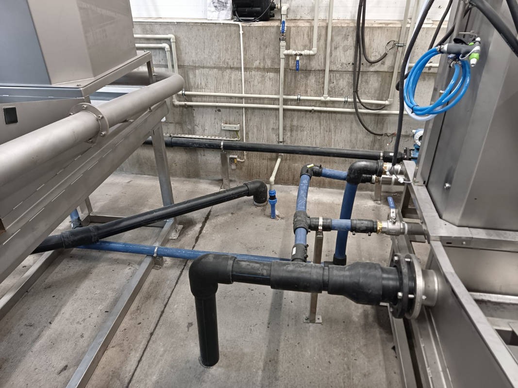 Instalacja przemysłowa z rurami z tworzyw sztucznych i elementami metalowymi, widoczne złącza, zawory i mocowania, na tle betonowej ściany z widocznymi rurami.