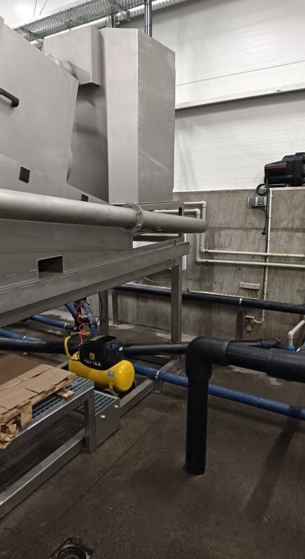 Fragment instalacji przemysłowej ze stali nierdzewnej z rurami, przewodami i żółtym kompresorem na podłodze w hali.