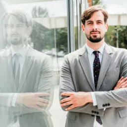 Młody mężczyzna w szarym garniturze i wzorzystym krawacie stoi z założonymi rękami na tle szklanej ściany, w której odbija się jego sylwetka.