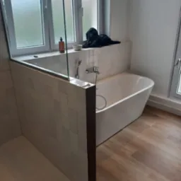 Wykończona łazienka z białą wanną wolnostojącą, kabiną prysznicową oddzieloną szklaną ścianką i drewnopodobną podłogą; na parapecie okiennym butelka z dozownikiem, miseczka i czarny ręcznik.