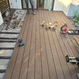 Estetik - Montaż desek na tarasie: widoczne częściowo ułożone deski kompozytowe, podkonstrukcja, narzędzia (wiertarki, przedłużacz, ukośnica) i osoba w jeansach. Część desek i gruzu pozostała po starym tarasie.