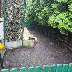 Estetik - Przygotowanie terenu pod przyszły ogród: wyrównana ziemia, widoczne drzewa iglaste wzdłuż ogrodzenia, fragment tarasu z drewnianymi krążkami i narzędzia ogrodnicze przy ścianie budynku.