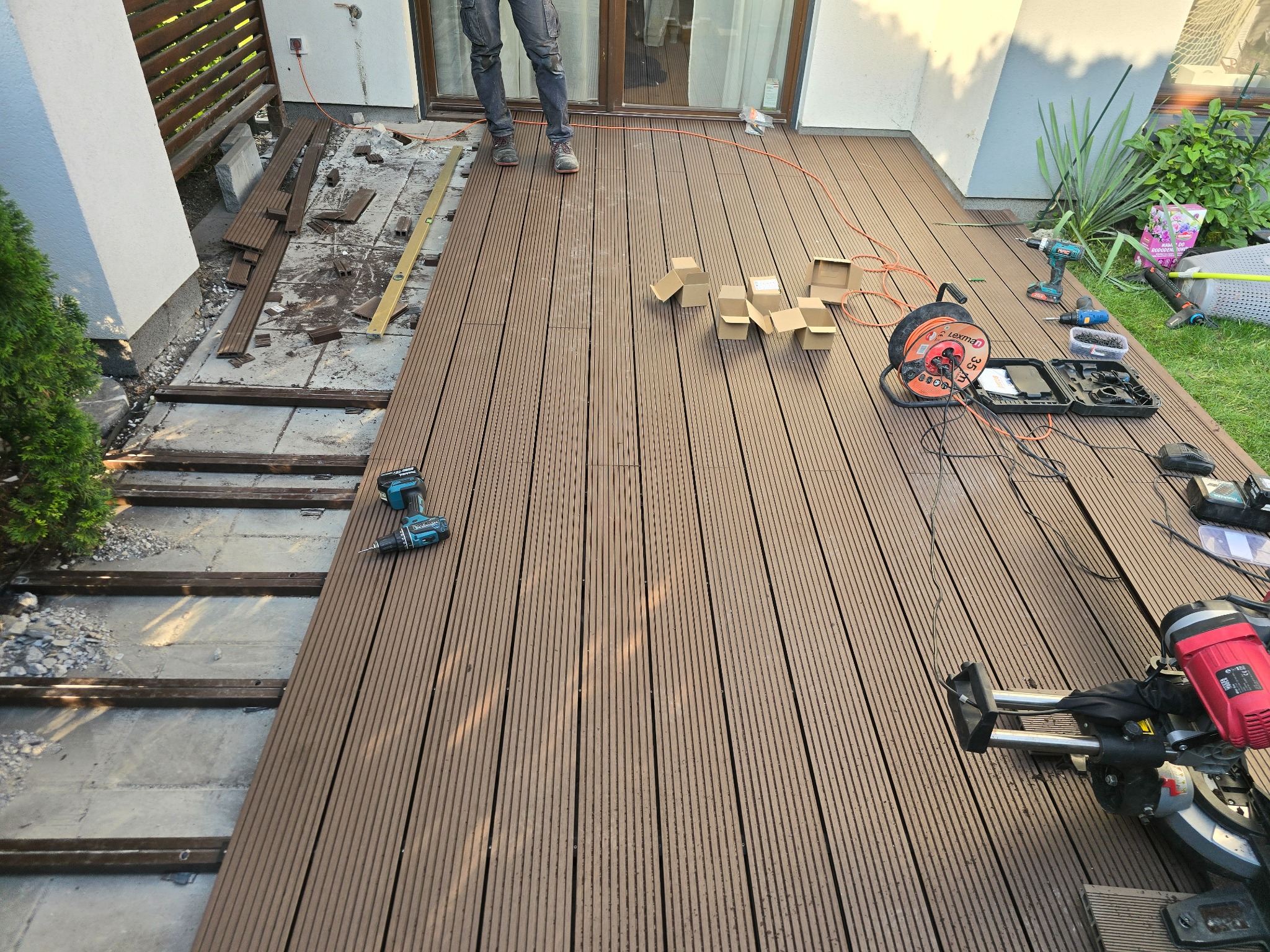 Montaż desek na tarasie: widoczne częściowo ułożone deski kompozytowe, podkonstrukcja, narzędzia (wiertarki, przedłużacz, ukośnica) i osoba w jeansach. Część desek i gruzu pozostała po starym tarasie.