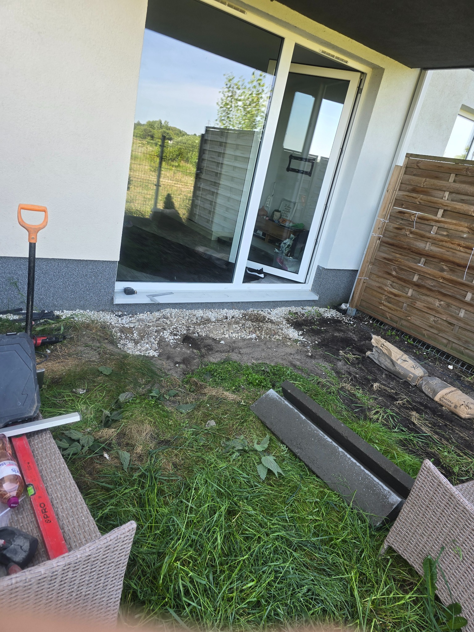 Przygotowania do montażu tarasu: widoczny fragment podłoża z kamykami, narzędzia (szpadel, poziomica), elementy konstrukcyjne tarasu (obrzeża) oraz worki z zaprawą murarską przy ścianie budynku...