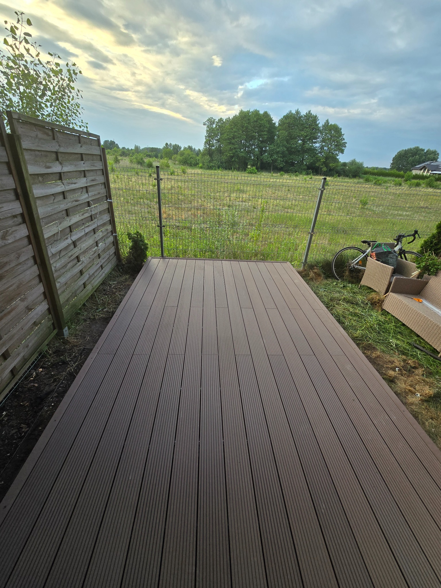 Nowo ułożony brązowy taras z desek kompozytowych, widok z góry na ogród z siatką ogrodzeniową, rowerem i częściowo rozpakowanymi meblami ogrodowymi.