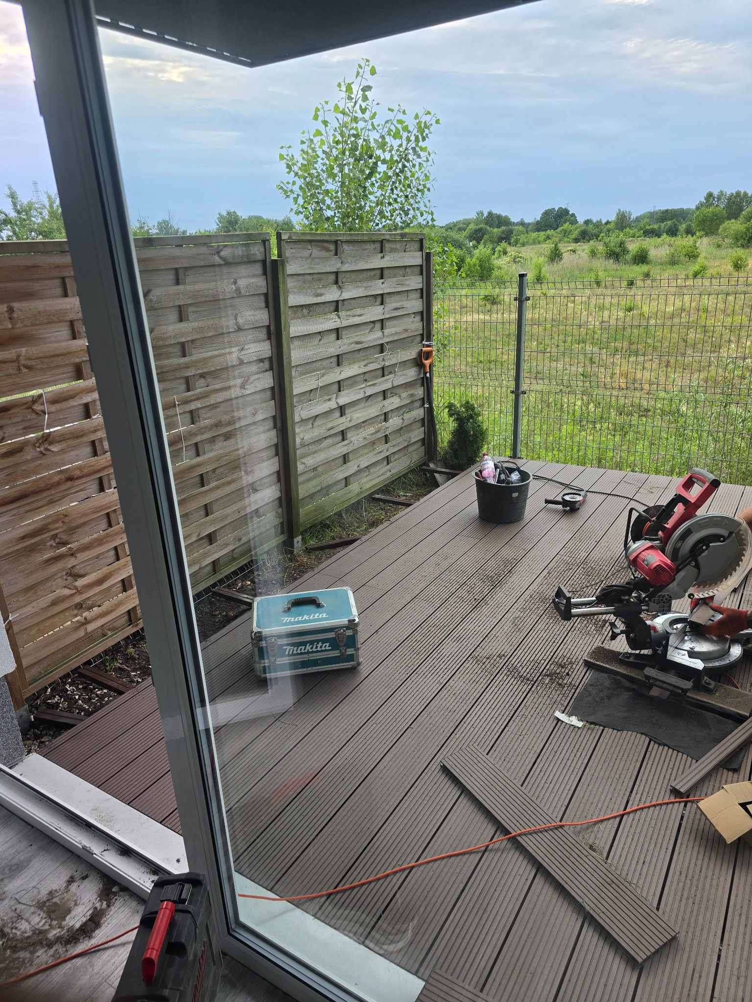 Montaż tarasu kompozytowego: widok z wnętrza domu na częściowo ułożony taras z desek kompozytowych, narzędzia pracy (ukośnica, szlifierka kątowa, skrzynka narzędziowa Makita) i elementy...