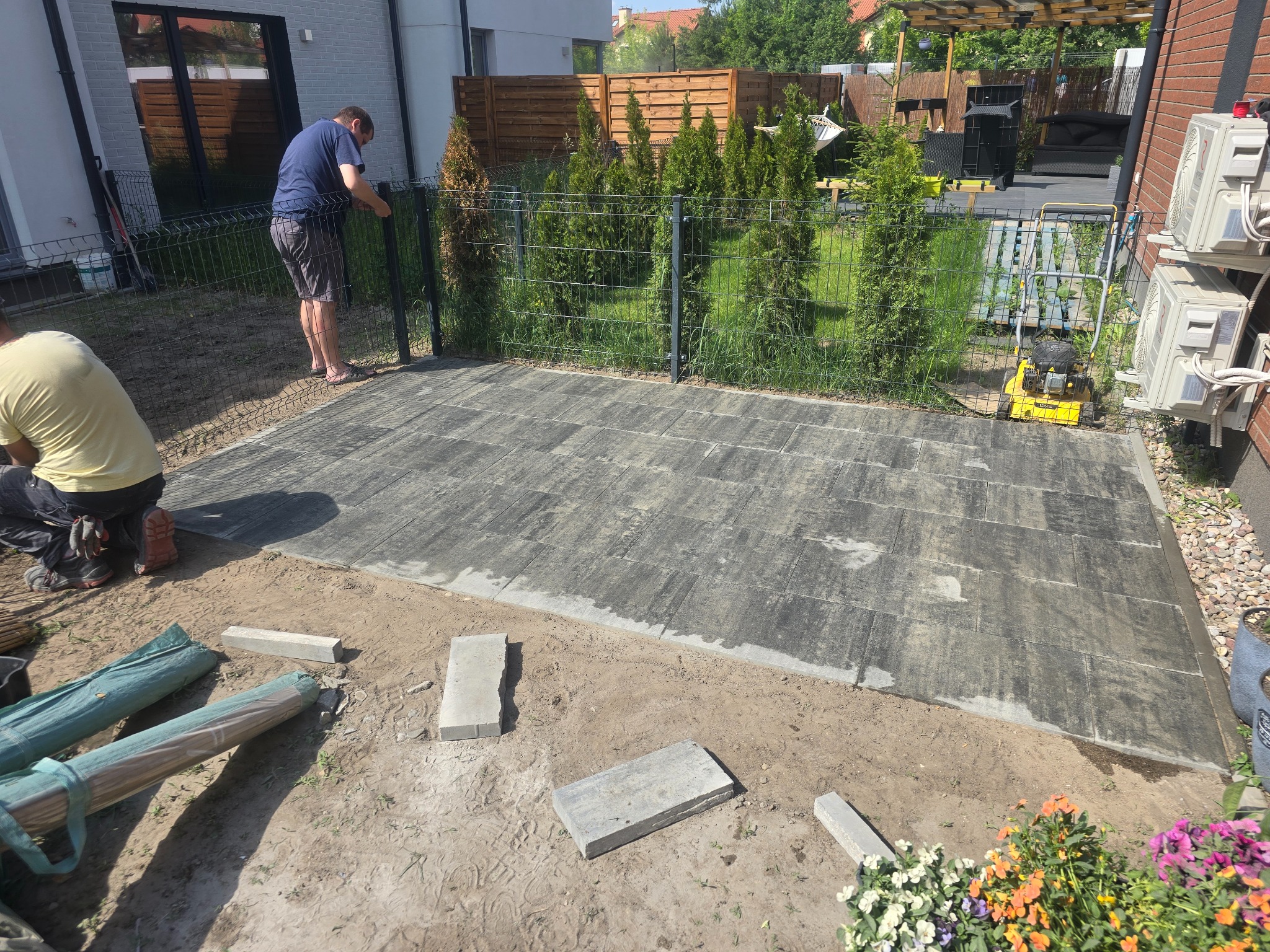 Układanie szarej kostki brukowej na przygotowanym podłożu w ogrodzie, widoczne narzędzia i materiały budowlane, pracownicy w trakcie pracy, trawnik i tuje w tle.