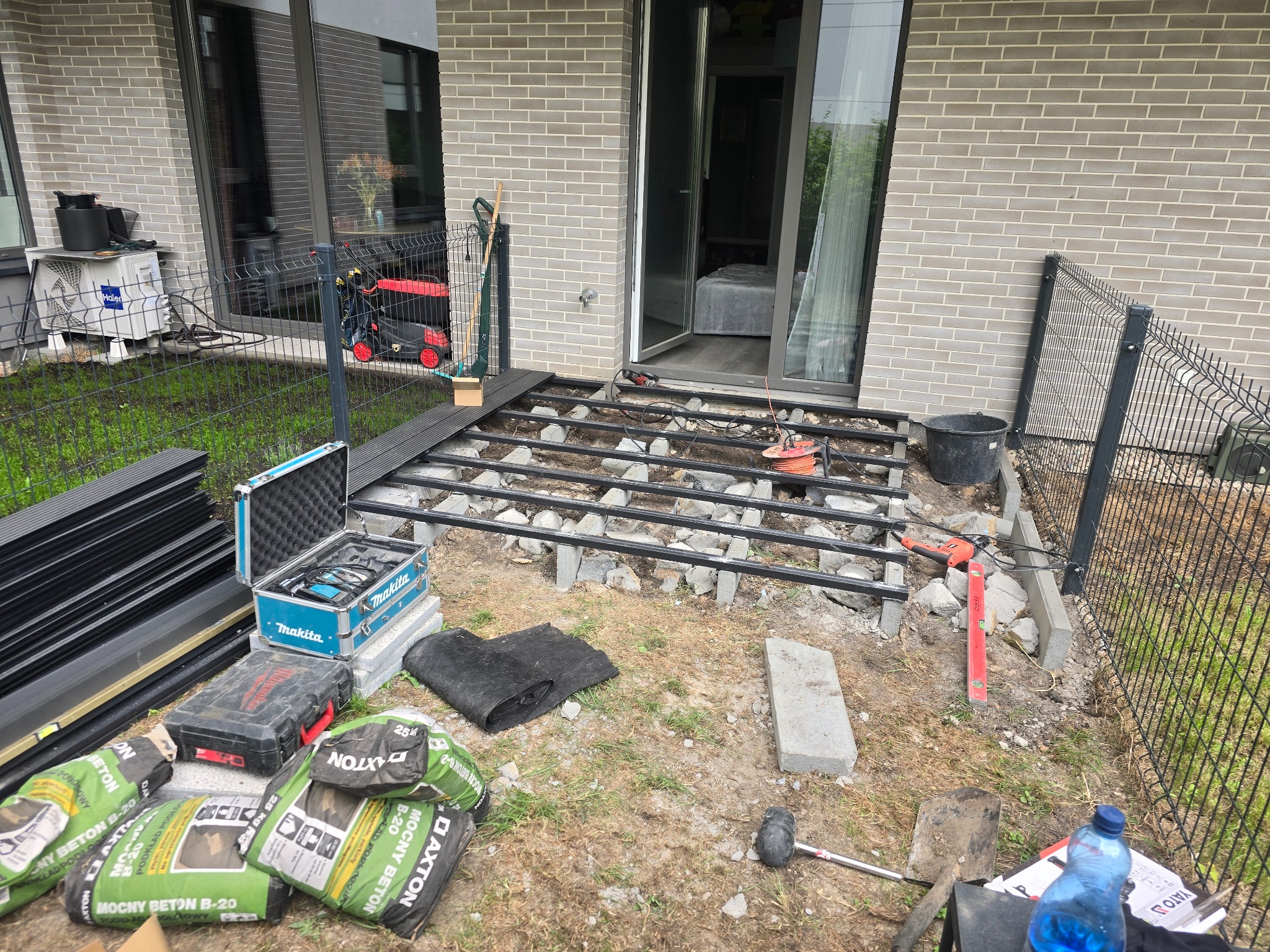 Etap montażu podkonstrukcji tarasu z profili stalowych na betonowych bloczkach, widoczne worki z suchym betonem, narzędzia oraz płyty kompozytowe w tle.