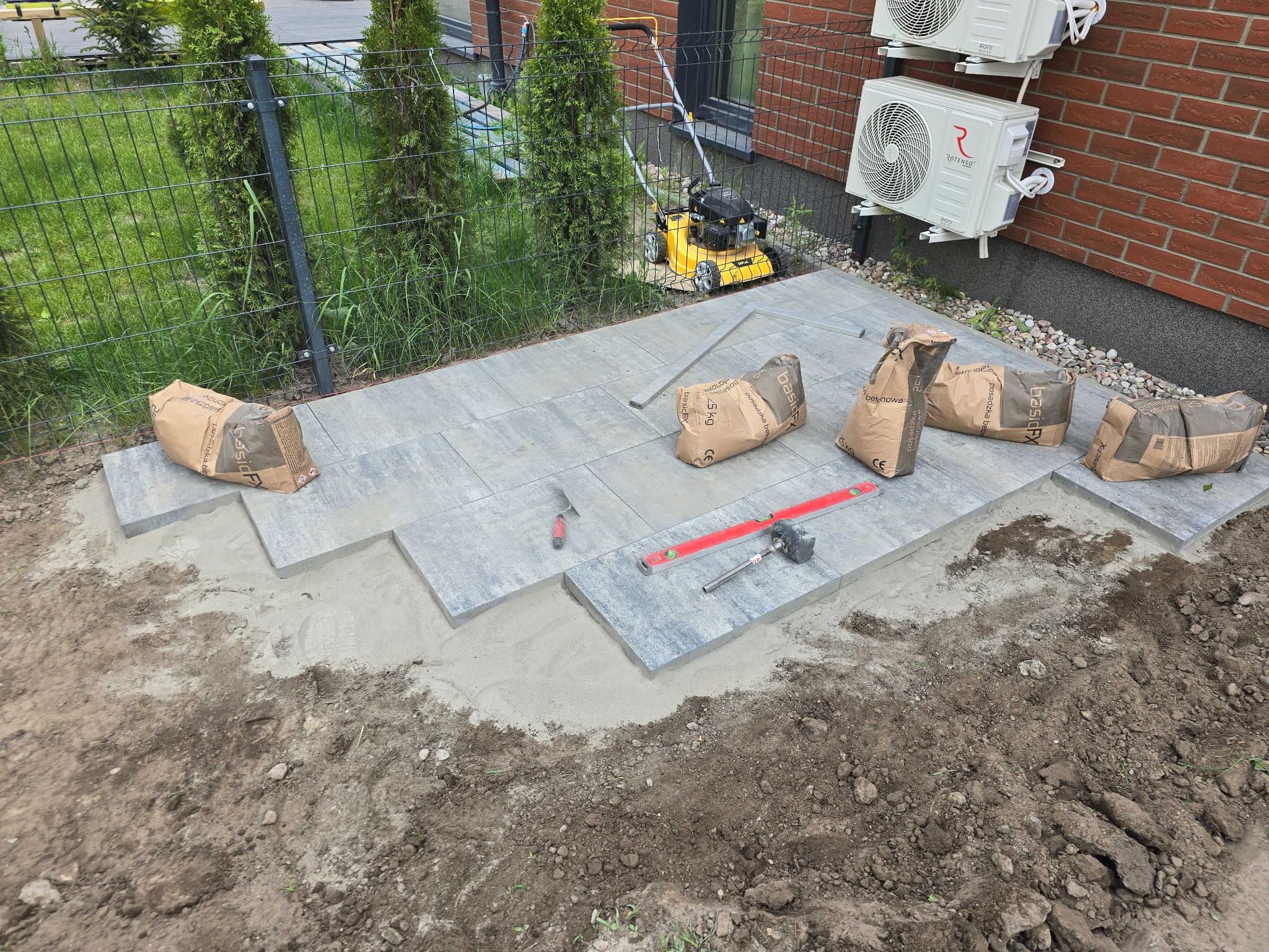 Układanie szarej kostki brukowej na podsypce piaskowej, widoczne narzędzia: poziomica, kielnia, młotek gumowy oraz worki z zaprawą, w tle trawnik, żywopłot i budynek z klimatyzacją.