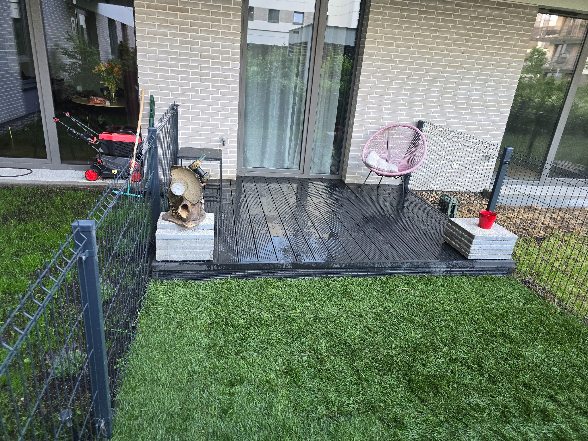 Nowoczesny taras z ciemnych desek kompozytowych, otoczony sztuczną trawą i ogrodzeniem z siatki, z widocznym krzesłem w stylu boho i elementami dekoracyjnymi.