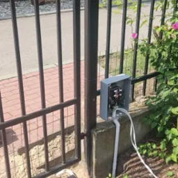 Instalacja domofonu przy metalowej bramie wjazdowej, widoczne okablowanie i puszka sterująca, ziemia wokół w trakcie prac ziemnych.