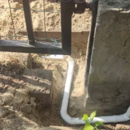 Biała rura karbowana ułożona w ziemi obok betonowego słupka i metalowej furtki, prawdopodobnie instalacja elektryczna lub telekomunikacyjna pod ziemią.