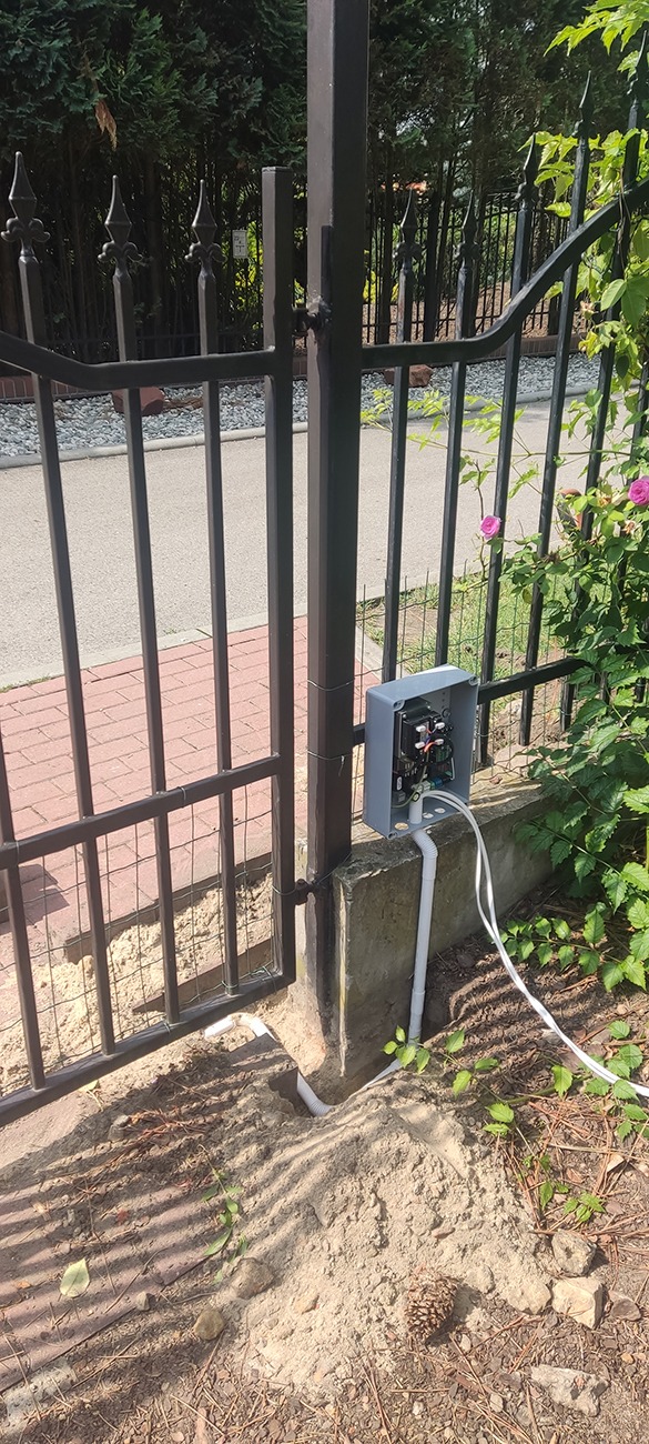 Instalacja domofonu przy metalowej bramie wjazdowej, widoczne okablowanie i puszka sterująca, ziemia wokół w trakcie prac ziemnych.