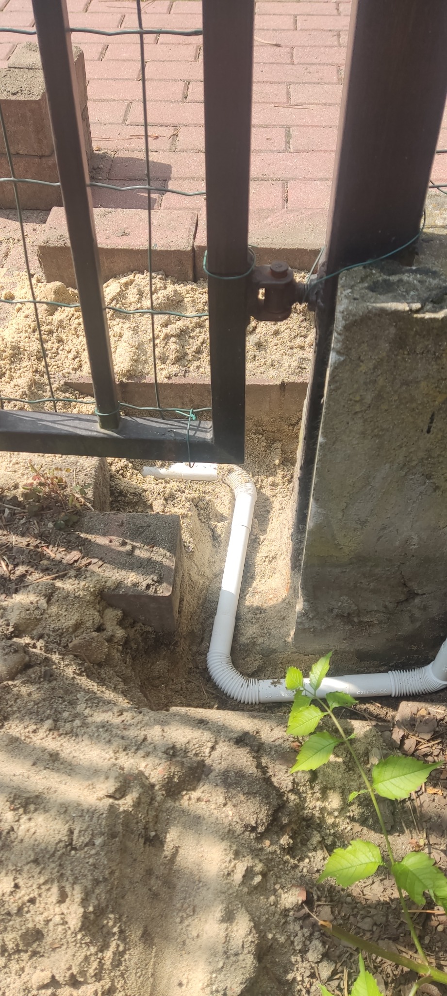 Biała rura karbowana ułożona w ziemi obok betonowego słupka i metalowej furtki, prawdopodobnie instalacja elektryczna lub telekomunikacyjna pod ziemią.
