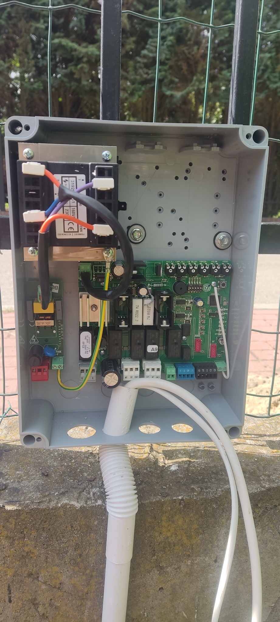 Otwarta szara skrzynka sterująca z widocznymi elementami elektronicznymi, w tym transformatorem, płytką drukowaną i złączami przewodów, zamontowana na zewnątrz na tle ogrodzenia i drzew.