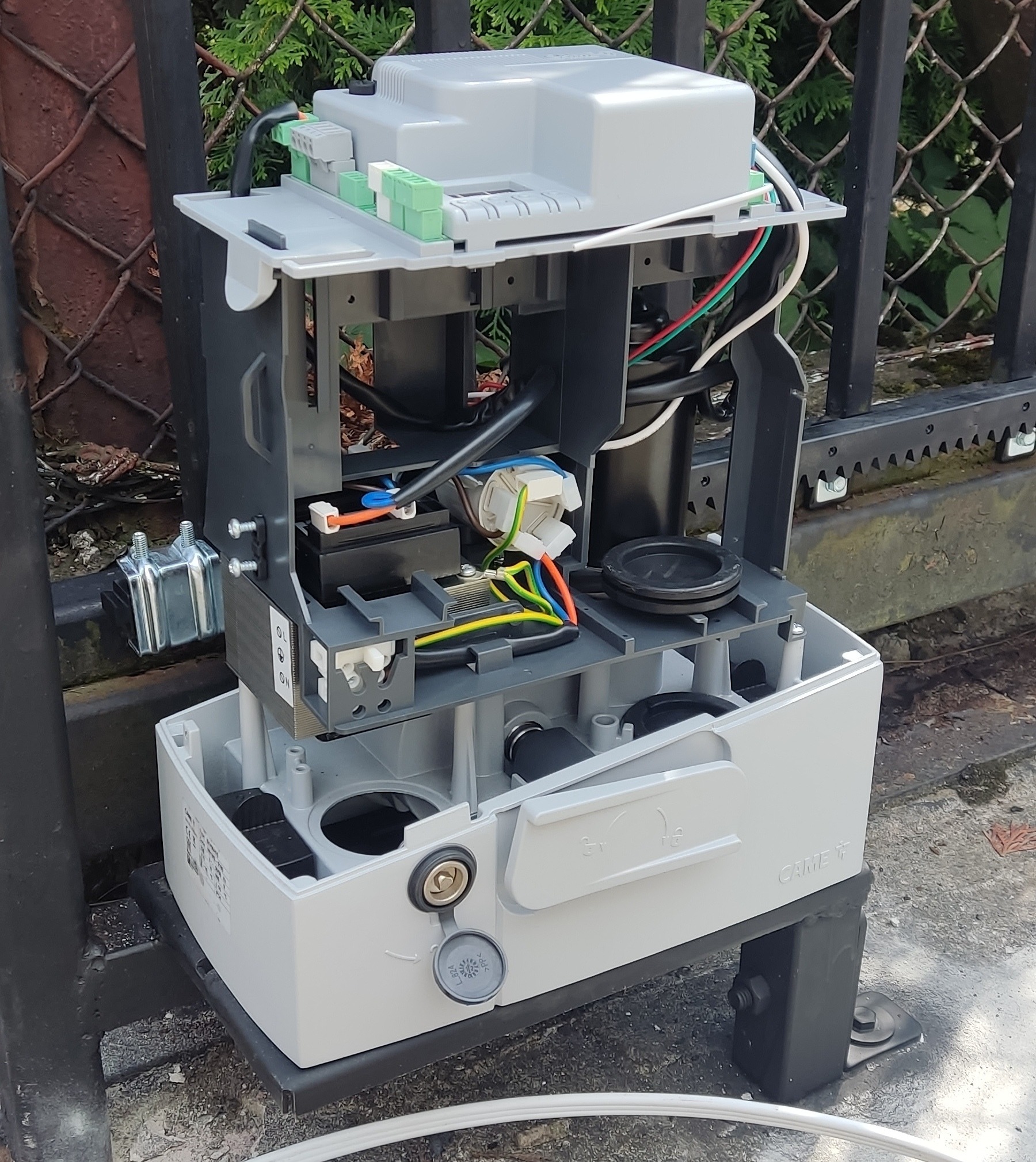 Otwarta obudowa automatyki bramy przesuwnej marki CAME, widoczne okablowanie, akumulator i podzespoły elektroniczne, zamontowana na słupku ogrodzenia.