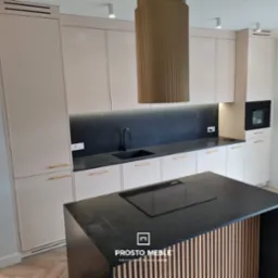 Nowoczesna kuchnia z wyspą, beżowe szafki z delikatnymi złotymi uchwytami, czarny blat i backsplash z subtelnym oświetleniem LED, oraz dekoracyjny okap w kształcie walca.