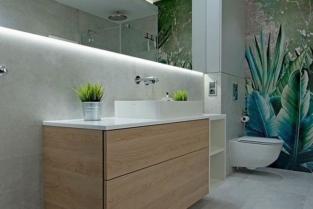 Nowoczesna łazienka w Poznaniu z drewnianą szafką pod umywalką, białym blatem i umywalką w kształcie prostokąta, podświetlanym lustrem oraz tapetą z motywem tropikalnych liści w tle.