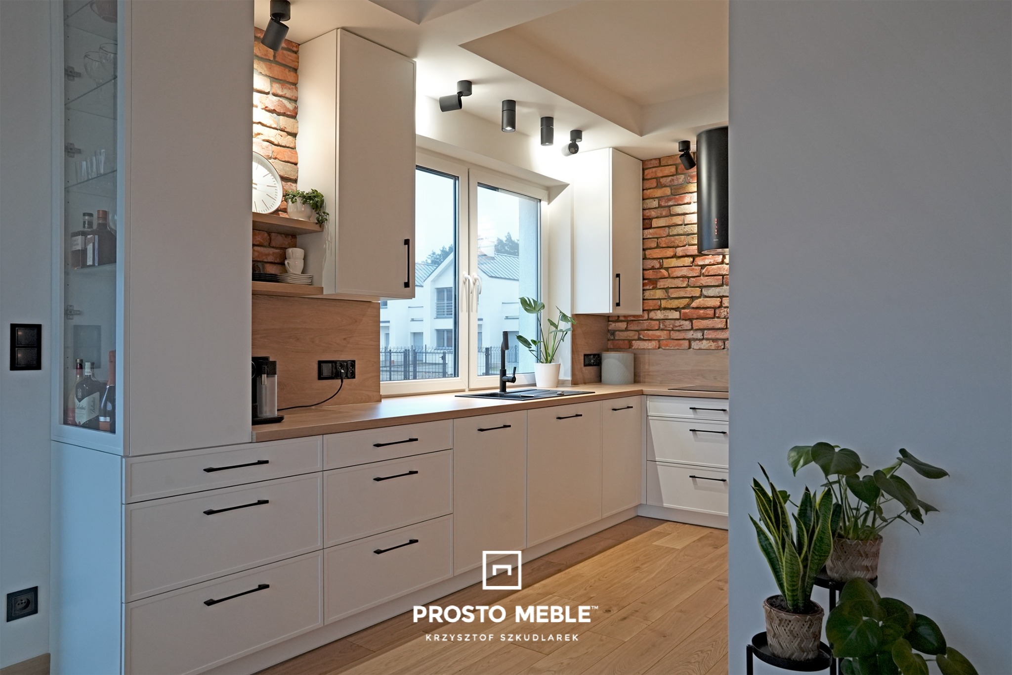 Jasna kuchnia z białymi frontami, drewnianym blatem i ceglaną ścianą, oświetlona punktowymi lampami sufitowymi, z widocznymi roślinami w doniczkach na parapecie i obok ściany.