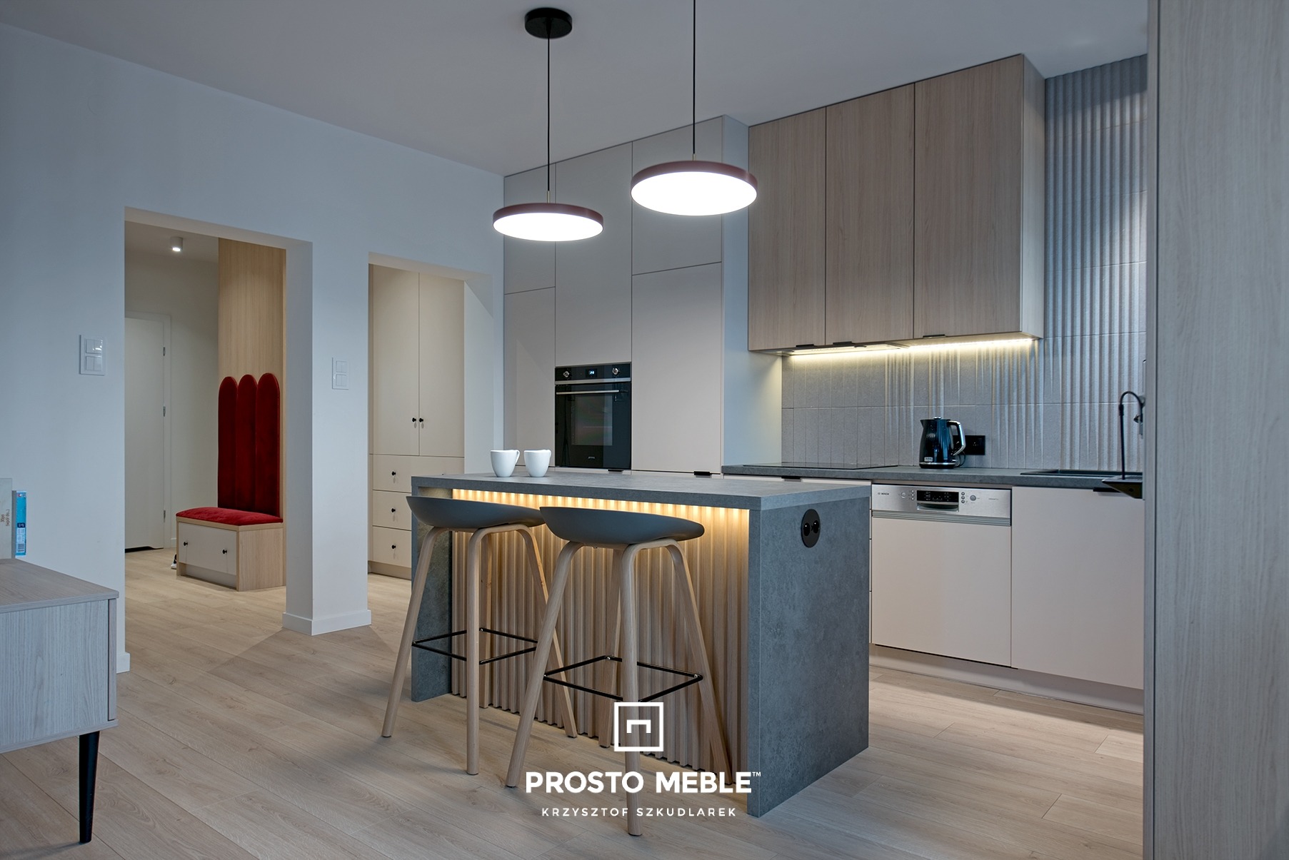Nowoczesna kuchnia z wyspą oświetloną od dołu, z szarymi stołkami barowymi i drewnianymi frontami szafek, widok na przedpokój z oryginalnym siedziskiem z czerwonym oparciem.