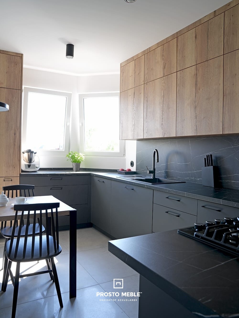 Nowoczesna kuchnia z szarymi szafkami dolnymi i drewnopodobnymi górnymi, czarnym blatem z marmurowym wzorem, widok na stół z czarnymi krzesłami i sprzęt AGD w tle.