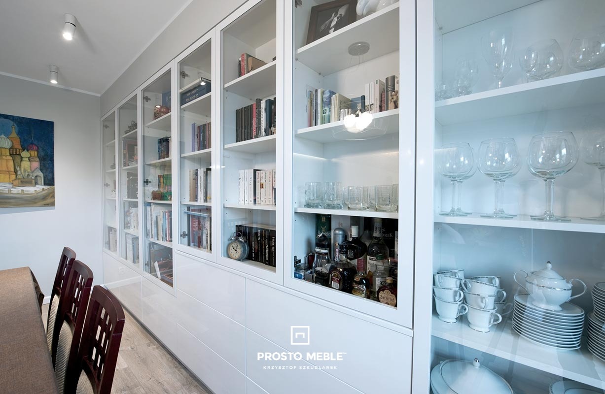 Biała zabudowa meblowa z przeszklonymi witrynami, eksponująca książki, zastawę stołową i alkohole, z logo firmy meblarskiej w dolnej części.