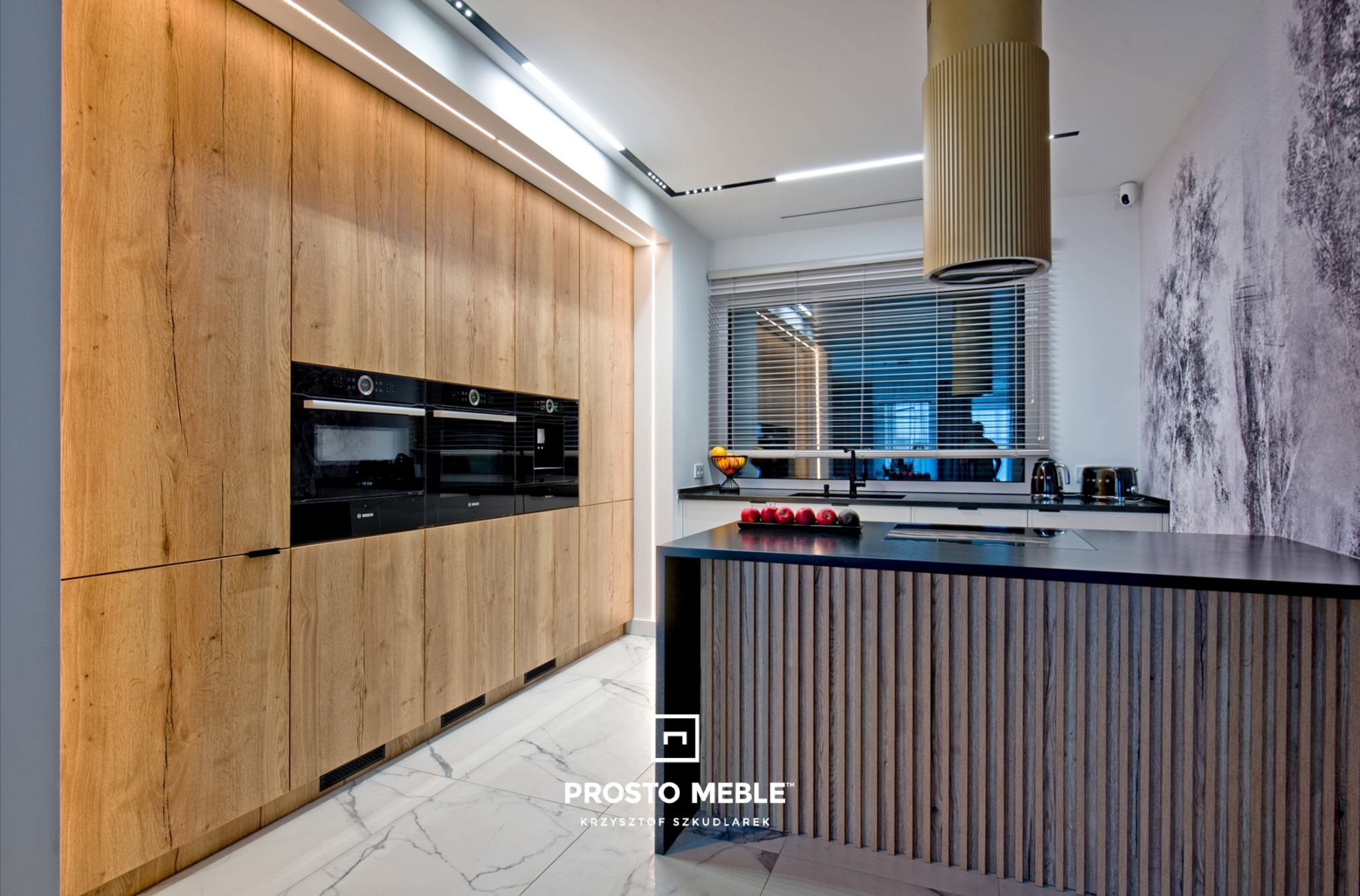 Nowoczesna kuchnia z drewnianymi frontami szafek, czarnymi piekarnikami w zabudowie i wyspą kuchenną z ryflowanym wykończeniem. Na blacie wyspy owoce w dekoracyjnej misie.