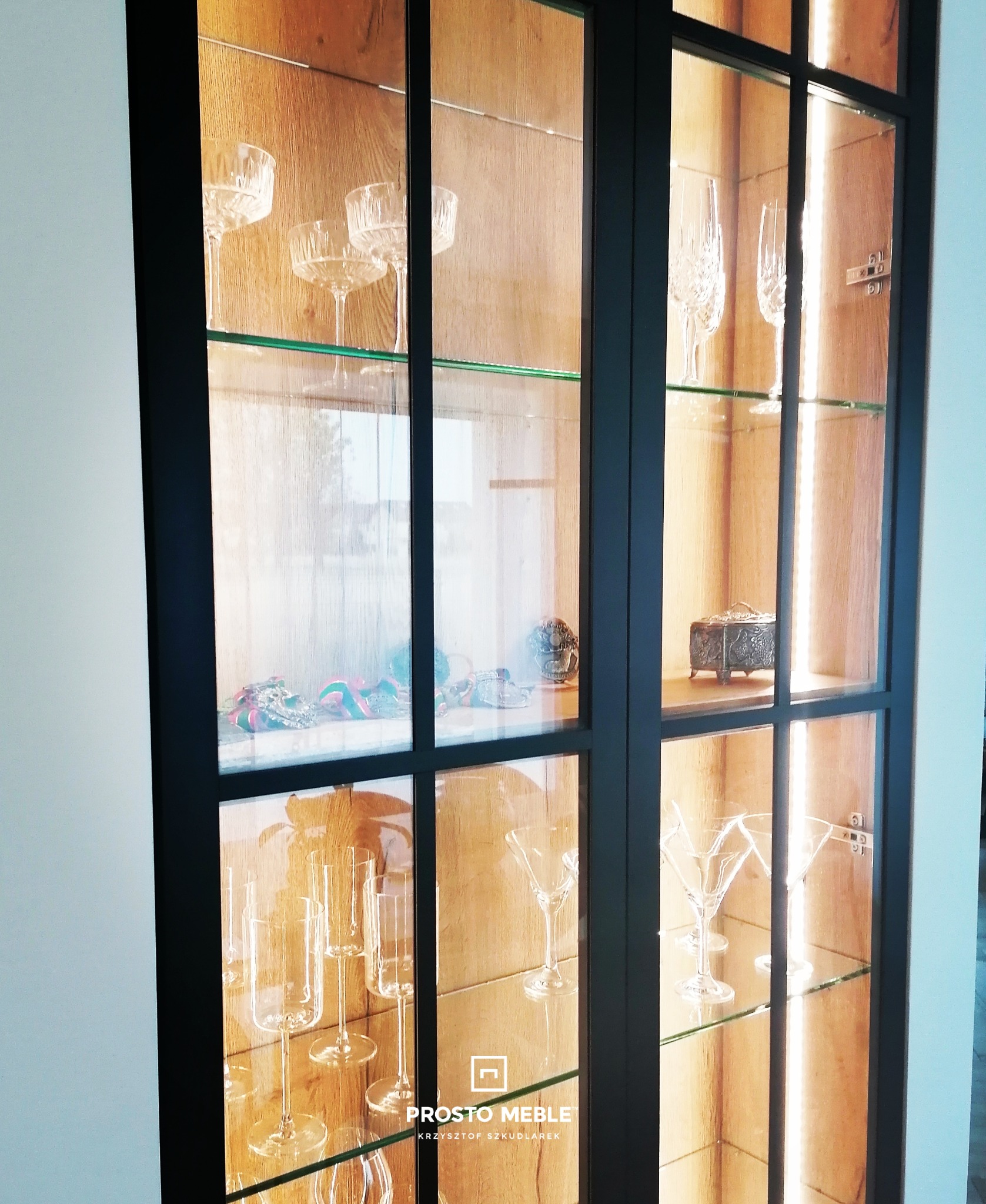 Nowoczesna witryna z czarnymi ramami, szklanymi półkami i oświetleniem LED, prezentująca kolekcję szkła dekoracyjnego i kryształów.