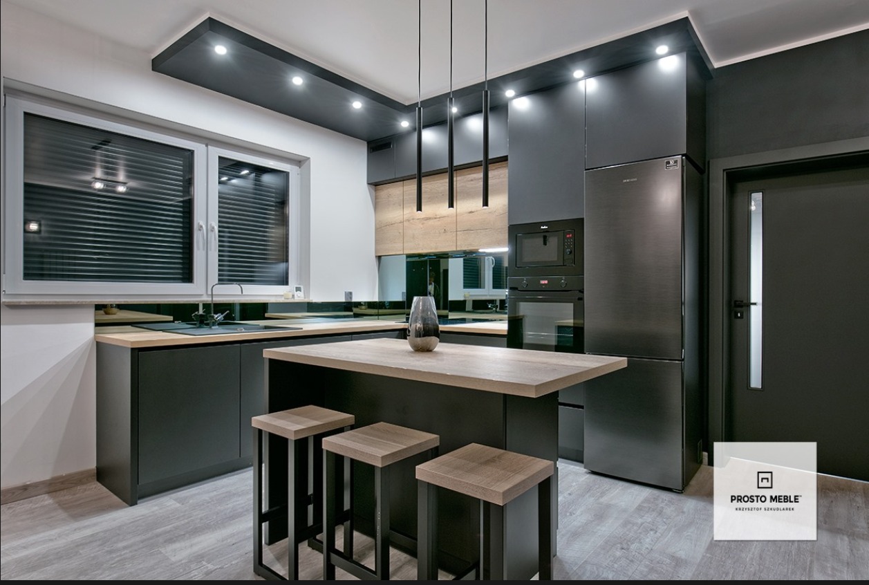 Nowoczesna kuchnia w odcieniach szarości i drewna z wyspą i trzema stołkami barowymi, oświetlona wiszącymi lampami, z widocznym oknem z żaluzjami i logo firmy meblarskiej w rogu.