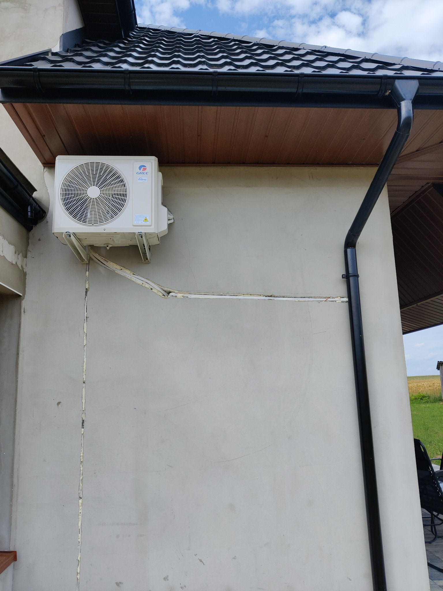 Zewnętrzna jednostka klimatyzacji marki Gree zamontowana na szarej ścianie budynku z widocznymi przewodami i rurami, czarna rynna dachowa w tle.