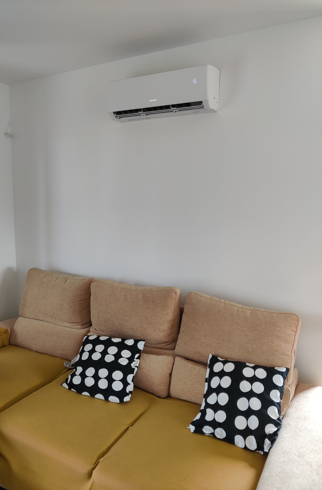 Biały klimatyzator marki Gree zamontowany na ścianie nad beżową kanapą z poduszkami w czarno-białe kropki. Na wyświetlaczu klimatyzatora widoczna temperatura 16 stopni.