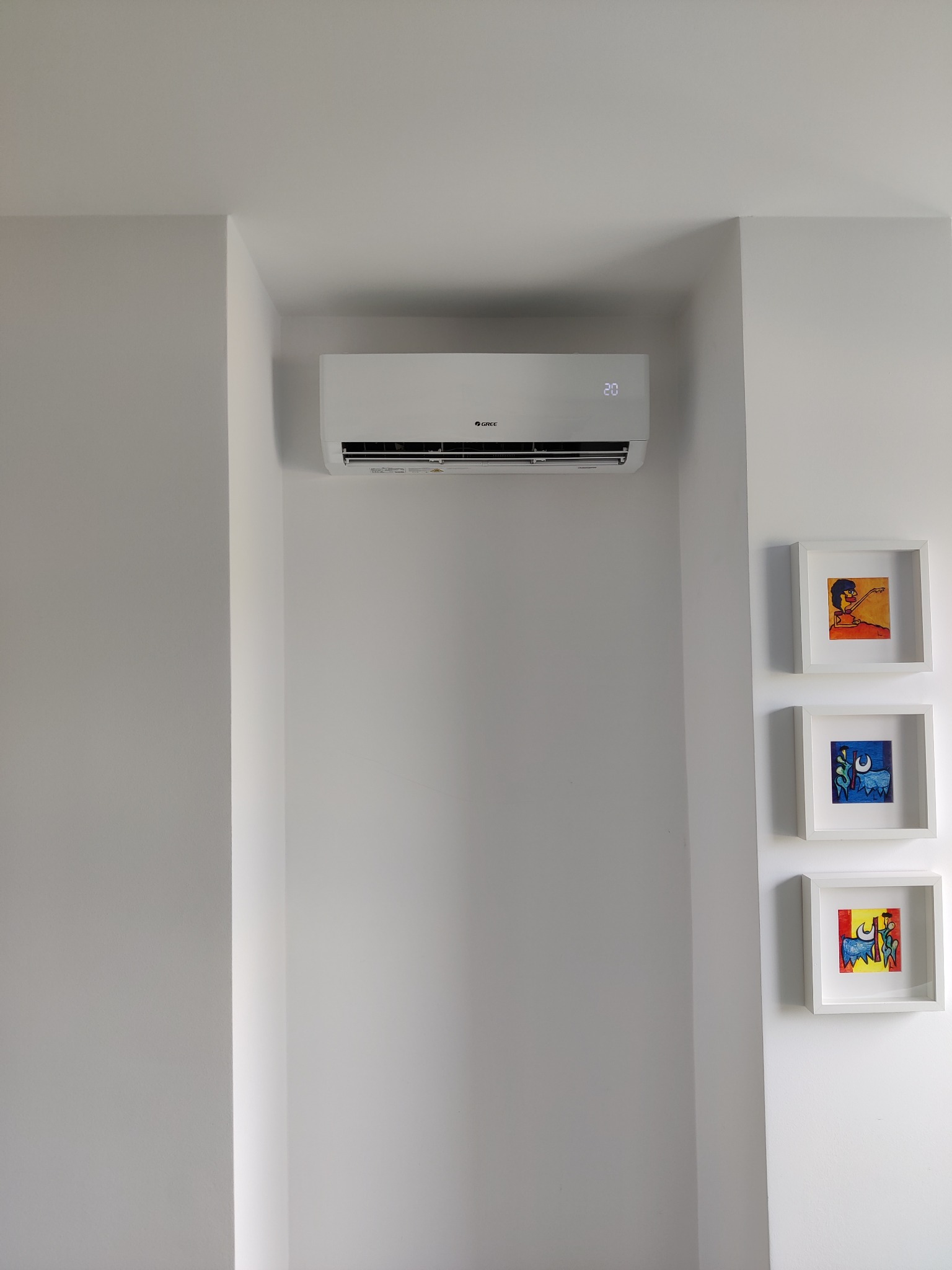 Biały klimatyzator marki Gree zamontowany na białej ścianie w minimalistycznym wnętrzu, obok trzy małe obrazy w białych ramkach.