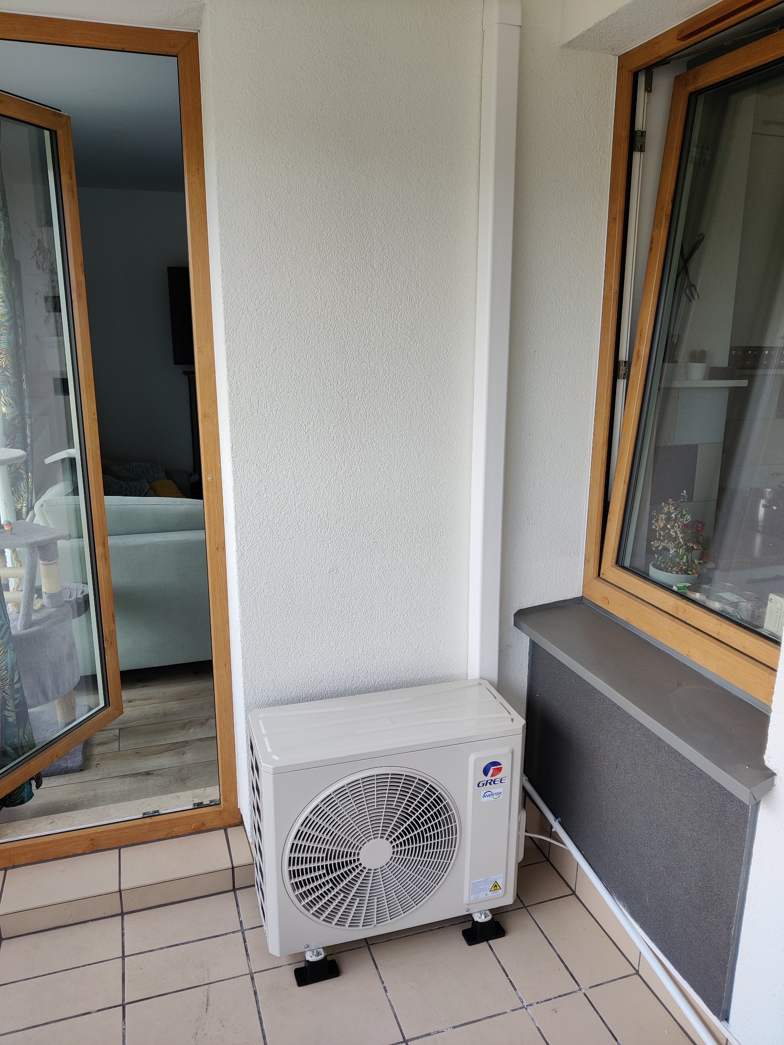 Zewnętrzna jednostka klimatyzacji GREE na balkonie z terakotową podłogą, widoczna z boku, z elementami montażowymi i rurami przy ścianie, obok drewnianej ramy okiennej i otwartych drzwi balkonowych.