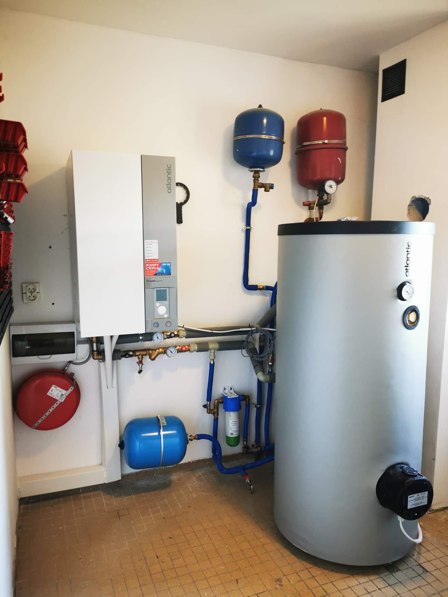 Kompletna instalacja grzewcza w pomieszczeniu technicznym: biały piec gazowy Atlantic, szary zasobnik ciepłej wody, czerwony i niebieski zbiornik wyrównawczy, filtr wody, liczne rury i zawory.