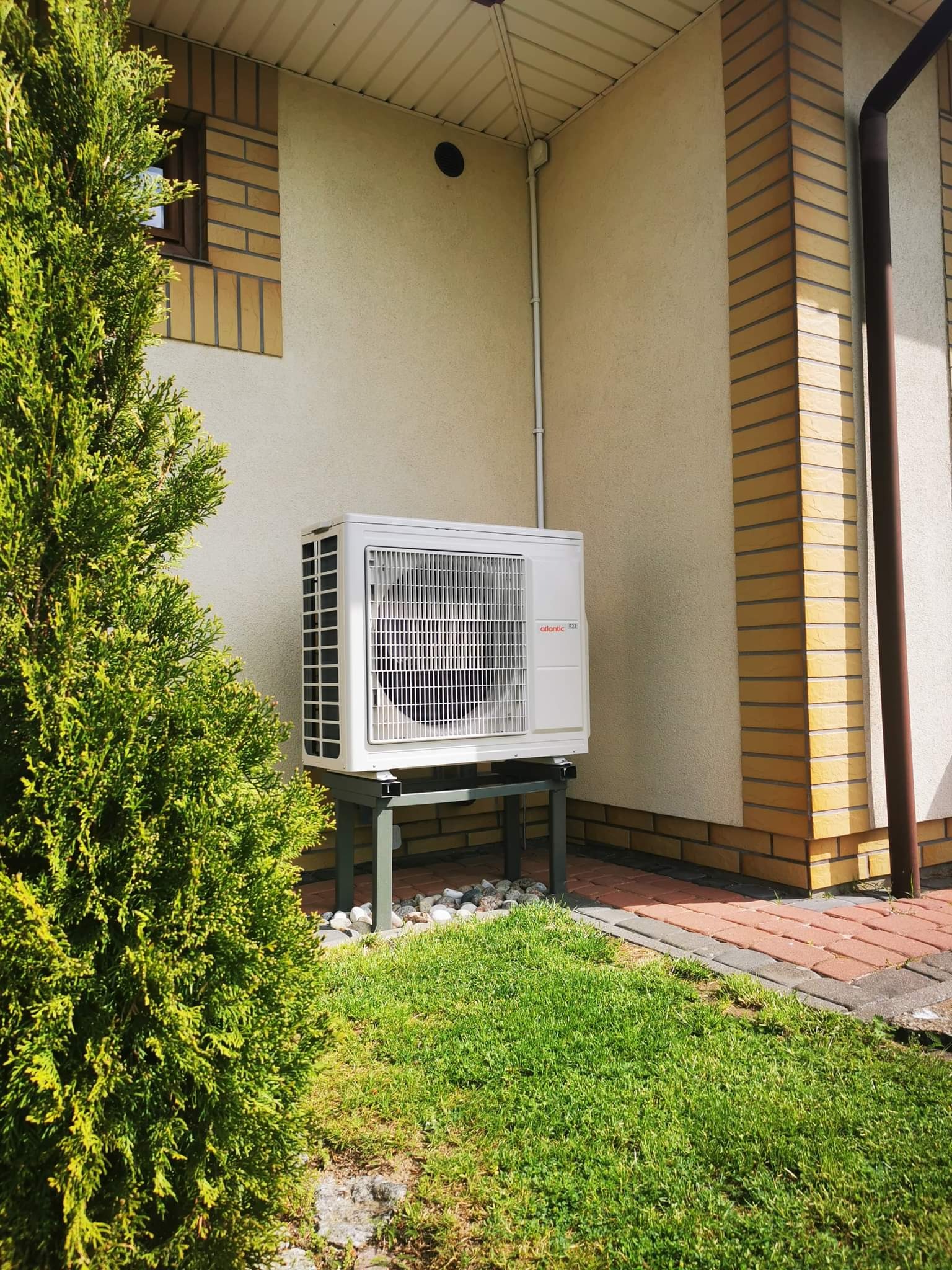 Biała jednostka zewnętrzna klimatyzacji Atlantic R32, zamontowana na metalowym stelażu na trawniku obok budynku z ceglaną elewacją i zielonym żywopłotem.