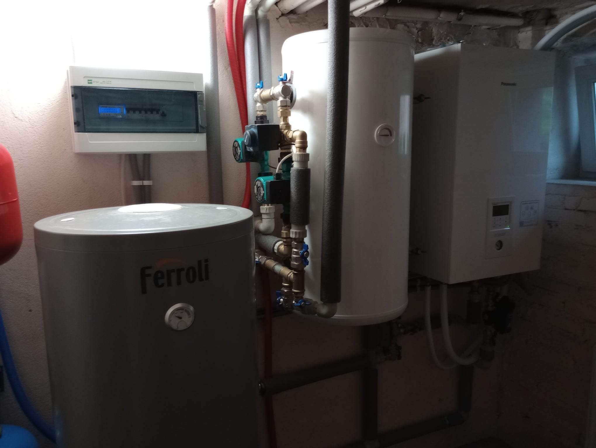 Kotłownia z szarym zbiornikiem Ferroli, białym zbiornikiem i białym kotłem Panasonic na tle ściany z instalacjami, widoczna skrzynka elektryczna.