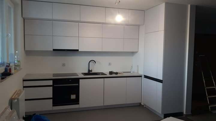 Nowoczesna, minimalistyczna kuchnia z białymi frontami szafek, czarną armaturą i sprzętem AGD, widoczna drabina w tle sugeruje prace wykończeniowe.