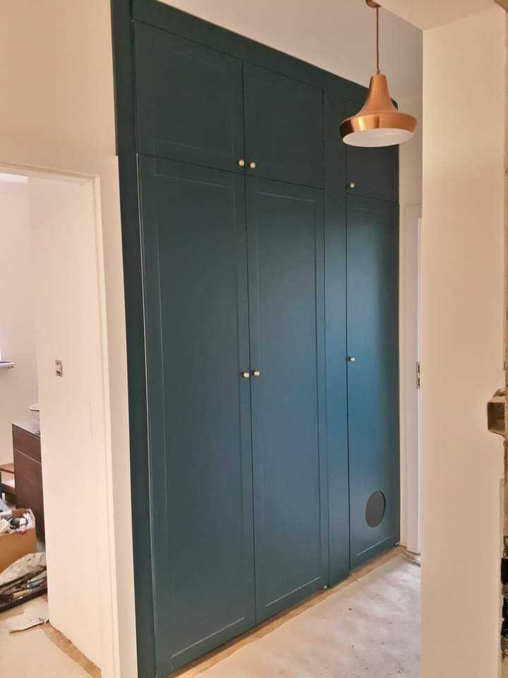 Wysoka, zabudowana szafa wnękowa w odcieniach ciemnej zieleni, z małymi, okrągłymi, złotymi uchwytami, umieszczona w korytarzu z widocznym wykończeniem podłogi i wiszącą miedzianą lampą.